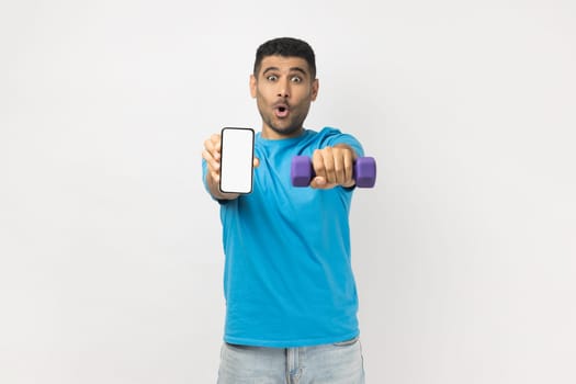 Portrait of surprised shocked man wearing T- shirt standing holding dumbbell and smartphone with empty display, advertisement for fitness app. Indoor studio shot isolated on gray background