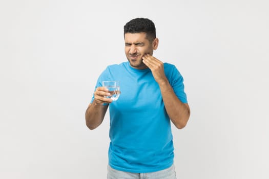 Portrait of unhealthy ill unshaven man wearing blue T- shirt standing with glass of cold water, feels pain in his tooth, has sensitive teeth. Indoor studio shot isolated on gray background.