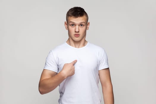 Portrait of surprised shocked teenager boy wearing T-shirt standing looking at camera with amazed expressing, pointing at himself with astonishment. Indoor studio shot isolated on gray background.