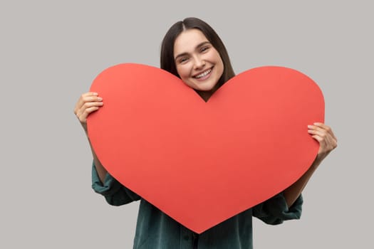 Portrait of happy charming woman holding of huge red heart, looking at camera, symbol of love, romantic emotions, wearing casual style jacket. Indoor studio shot isolated on gray background.