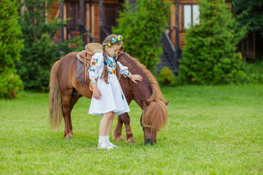 young girl in Ukrainian national dress strokes a pony that grazes on the lawn