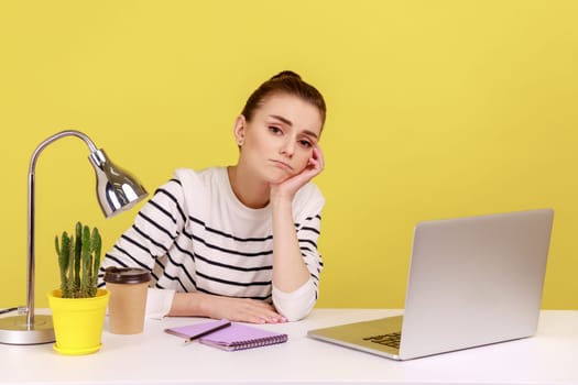 Bored unhappy woman manager sitting at workplace with laptop and leaning on hand, feeling lazy uninterested in her job, depression at work. Indoor studio studio shot isolated on yellow background.