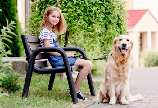 Preteen girl wearing jeans dress with golden retriever dog outdoors sitting on bench in summertime . Pretty kid with purebred fluffy doggy pet in city