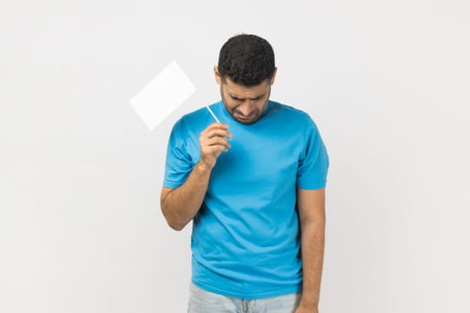 Portrait of depressed unshaven man wearing blue T- shirt standing with his head down holding a white flag, expressing sadness and sorrow. Indoor studio shot isolated on gray background.