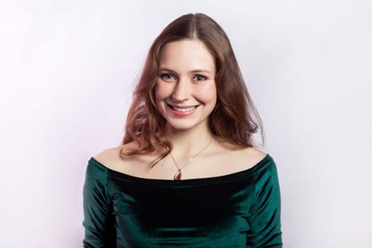 Portrait of smiling happy attractive woman wearing green dress standing looking at camera with toothy smile, expressing kind positive emotions. Indoor studio shot isolated on gray background.