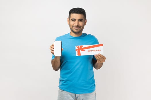 Portrait of happy satisfied man wearing T- shirt standing with gift voucher and showing smart phone with empty display, copy space for advertisement. Indoor studio shot isolated on gray background.