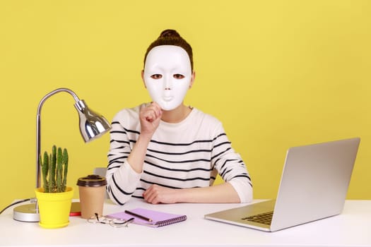 Portrait of unknown anonymous woman covering her face with white mask, hiding personality while sitting on workplace with laptop. Indoor studio studio shot isolated on yellow background.