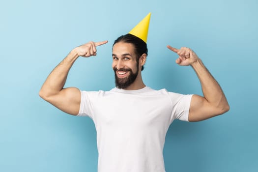 Portrait of satisfied festive man wearing white T-shirt celebrating his birthday, standing pointing at party cone on his head, smiling broadly. Indoor studio shot isolated on blue background.