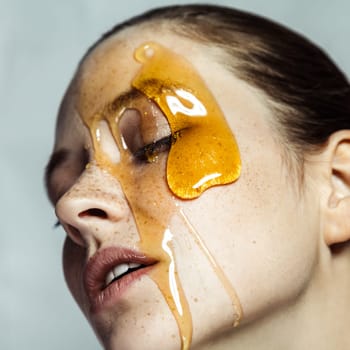 Closeup profile portrait of attractive young brunette woman with freckles, making honey homemade mask for skincare, posing with closed eyes. Indoor studio shot isolated on gray background.