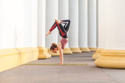 Full length side view portrait of young adult sporty attractive woman in sporty pants and top practicing yoga outdoor, standing in crane, bakasana pose, working out, healthy lifestyle.
