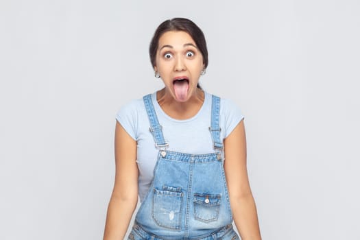 Portrait of disobedient crazy woman wearing denim overalls sticking out tongue and grimacing, aping showing derisive, naughty expression. Indoor studio shot isolated on gray background.