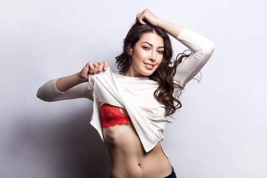 Portrait of attractive smiling woman with wavy hair taking off her shirt, raised her arms, showing her naked belly and red lingerie. Indoor shot isolated on gray background.