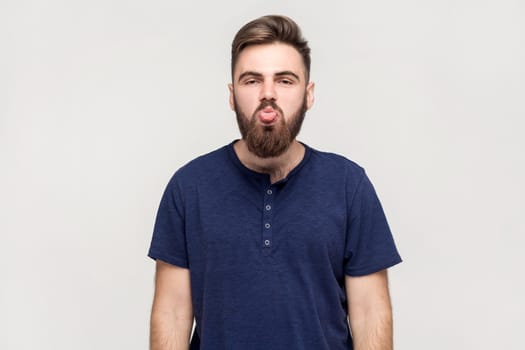 Portrait of man with beard wearing dark blue T-shirt showing out tongue and looking at camera with naughty disobedient grimace, making face. Indoor shot isolated on gray background.