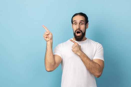 Portrait of surprised man with beard in white T-shirt pointing finger aside, showing copy space for commercial text, blank wall with idea presentation. Indoor studio shot isolated on blue background.