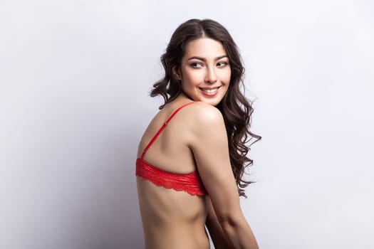 Side view portrait of smiling satisfied shy woman sensuality model with curly hair looking away with smile, hiding her red lingerie. Indoor studio shot isolated on gray background.