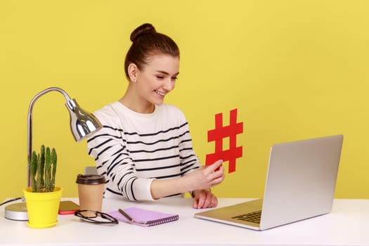 Satisfied woman sitting at workplace, showing big red hashtag symbol to laptop screen, sharing viral content, tagged message. Indoor studio studio shot isolated on yellow background.