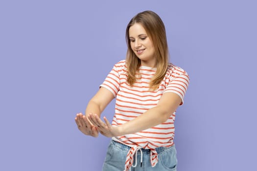 Portrait of satisfied happy blond woman wearing striped T-shirt looking at her nails with toothy smile, admires her freshly painted nails. Indoor studio shot isolated on purple background.