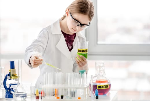 Smart girl doing scientific chemistry experiment wearing protection glasses, and add liquid to bottle with title danger. Schoolgirl with chemical equipment on school lesson