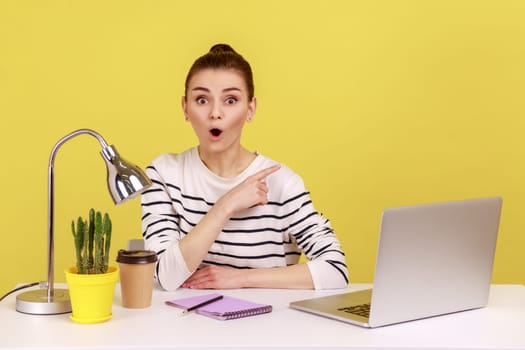 Look at advertisement. Shocked young woman sitting at office workplace with laptop and pointing aside, showing copy space for promotional text. Indoor studio studio shot isolated on yellow background