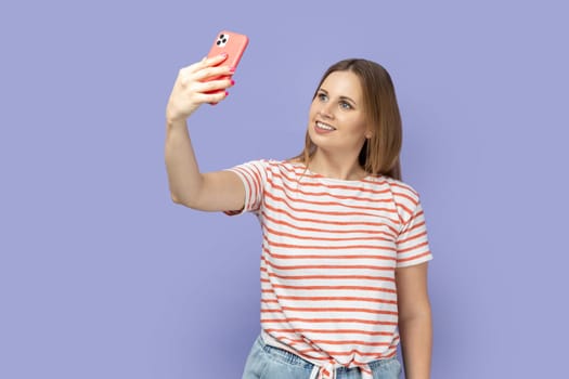 Portrait of smiling blond woman in striped T-shirt standing with phone and having video call or broadcasting livestream, looking smiling at screen. Indoor studio shot isolated on purple background.