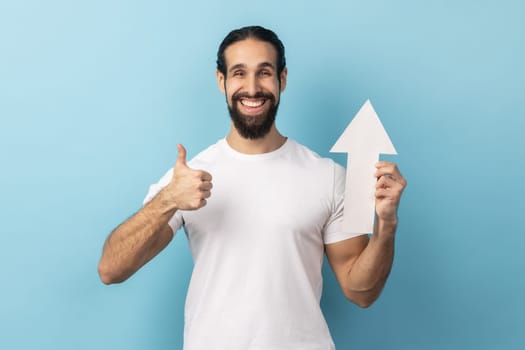 Portrait of happy handsome man with beard wearing white T-shirt showing white arrow pointing up, showing thumb up, like gesture, looking at camera. Indoor studio shot isolated on blue background.