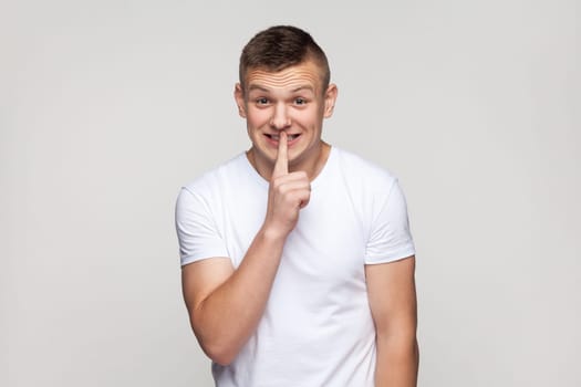 Portrait of happy attractive teenager boy wearing T-shirt looking away holding finger near lips, keeping secret, expressing positive emotions. Indoor studio shot isolated on gray background.