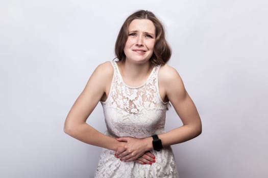 Portrait of sick ill beautiful woman wearing white dress holding her belly, having terrible period pain, feels discomfort, frowning her face. Indoor studio shot isolated on gray background.