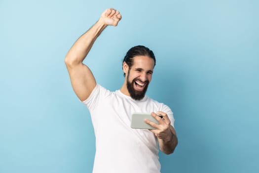 Portrait of man with beard wearing white T-shirt holding mobile phone showing yes gesture sincerely rejoicing, victory in online virtual game. Indoor studio shot isolated on blue background.