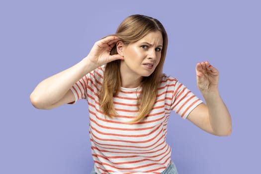 Portrait of blond woman wearing striped T-shirt holding hand near ear trying to listen quiet conversation, overhearing gossip, showing blah gesture. Indoor studio shot isolated on purple background.