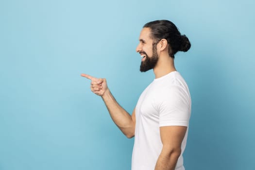 Side view of smiling man with beard in white T-shirt pointing finger aside, showing copy space for commercial text, blank wall with idea presentation. Indoor studio shot isolated on blue background.