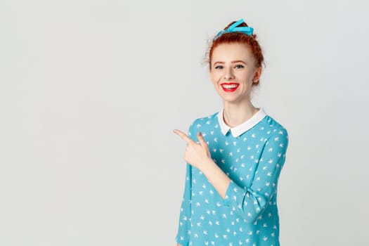 Portrait of smiling delighted ginger woman with bun hairstyle, pointing aside at advertisement area, copy space for promotion, wearing blue dress. Indoor studio shot isolated on gray background.