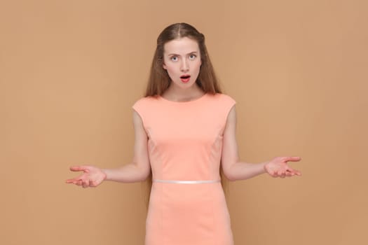 Portrait of angry shocked upset woman with long hair standing with raised arms, asking what, looking at camera, wearing elegant dress. Indoor studio shot isolated on brown background.