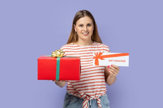 Portrait of joyful cheerful blond woman wearing striped T-shirt holding gift voucher and red gift box, preparing present for giveaway. Indoor studio shot isolated on purple background.