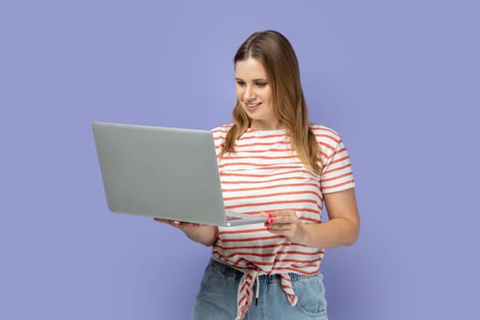 Portrait of smiling positive blond woman wearing striped T-shirt working on laptop, looking at display, enjoying her online work, reading information. Indoor studio shot isolated on purple background.