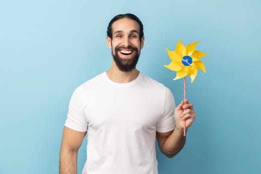 Portrait of cheerful childish man with beard wearing white T-shirt holding paper windmill, pinwheel toy on stick, playing with paper toy. Indoor studio shot isolated on blue background.
