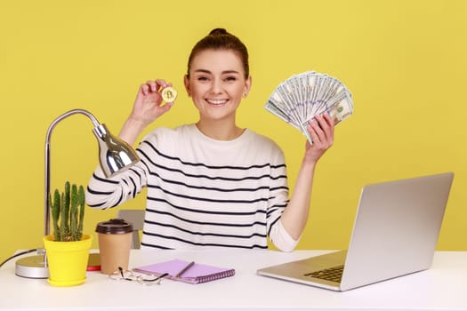 Satisfied woman office worker holding and showing fan of hundred dollar banknotes and bitcoin crypto coin looking at camera with toothy smile. Indoor studio studio shot isolated on yellow background.