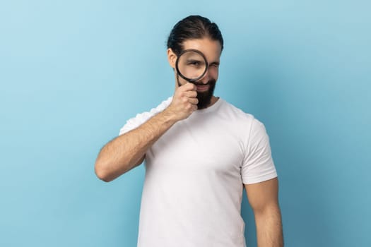 Portrait of man with beard wearing white T-shirt standing, holding magnifying glass and looking at camera with big zoom eye, verifying authenticity. Indoor studio shot isolated on blue background.