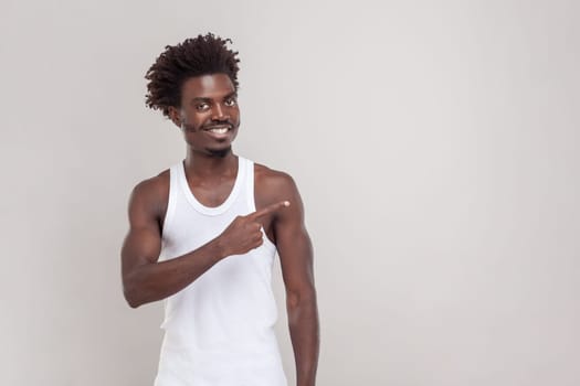 Portrait of satisfied delighted man with Afro hairstyle points away on copy space, discusses amazing promo, gives way or direction, wearing T-shirt. Indoor studio shot isolated on gray background.