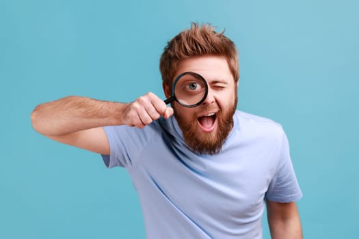 Portrait of funny positive bearded man holding magnifying glass and looking at camera with big zoom eye, curious face, keeps mouth open. Indoor studio shot isolated on blue background.