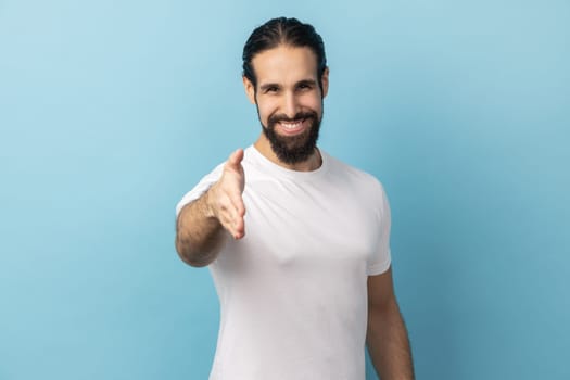 Portrait of man with beard wearing white T-shirt standing and looking at camera with toothy smile and giving hand to greeting or handshake. Indoor studio shot isolated on blue background.