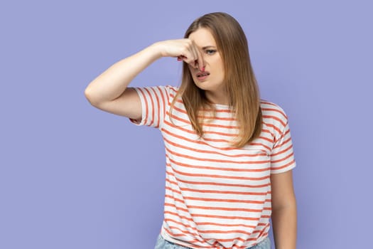 Portrait of unhappy attractive blond woman wearing striped T-shirt standing and pinching her nose, smelling, feels negative odor. Indoor studio shot isolated on purple background.