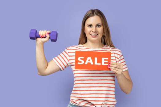 Charming smiling blond woman wearing striped T-shirt holding dumbbell in hand and showing sale card, advertising discounts for fitness workouts. Indoor studio shot isolated on purple background.