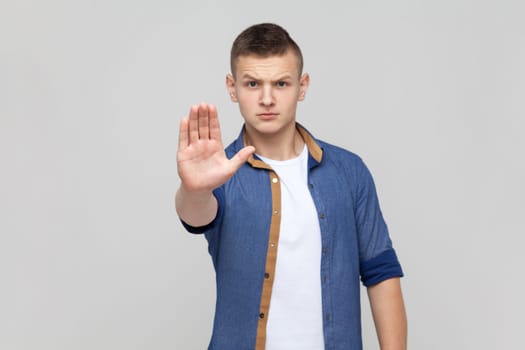 Portrait of teenager boy wearing blue shirt making stop gesture showing palm of hand, conflict prohibition warning about danger, stop bullying. Indoor studio shot isolated on gray background.