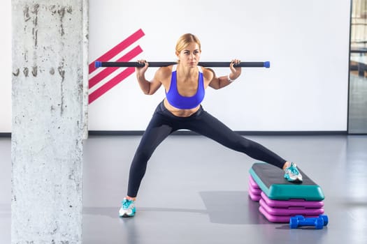 Portrait of attractive blonde sportswoman wearing tight sportswear with barbell on her shoulders doing side lunges on step platform, having sport workout in fitness club. Indoor shot.