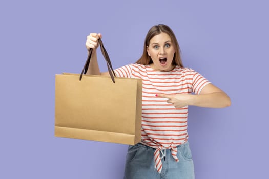 Portrait of astonished surprised blond woman wearing striped T-shirt standing pointing at shopping bag, being amazed of good purchase. Indoor studio shot isolated on purple background.