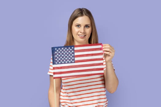 Portrait of blond woman wearing T-shirt holding in hand flag of united states of america, celebrating independence day, having calm facial expression. Indoor studio shot isolated on purple background.