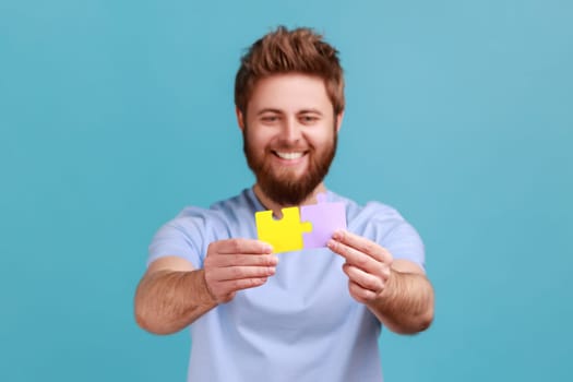 Portrait of positive happy bearded man combining to different colorful pieces of puzzle, making right solution, creating business association. Indoor studio shot isolated on blue background.