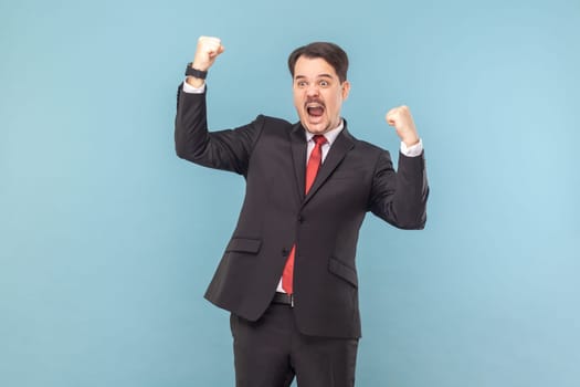 Portrait of excited extremely happy man standing with clenched fists, celebrating his victory, screaming hurray, wearing black suit with red tie. Indoor studio shot isolated on light blue background.