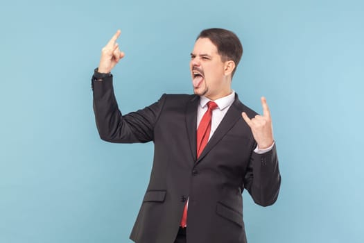 Portrait of crazy cool man with mustache standing showing rock and roll gesture, screaming with excitement, wearing black suit with red tie. Indoor studio shot isolated on light blue background.