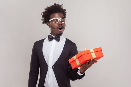 Portrait of surprised astonished man in glasses standing with red gift box, looking at present with open mouth, wearing white shirt and tuxedo. Indoor studio shot isolated on gray background.